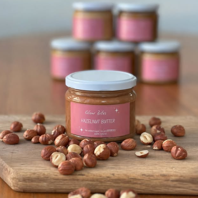 Jar of hazelnut butter on a wooden board with scattered nuts, surrounded by more jars in the background.