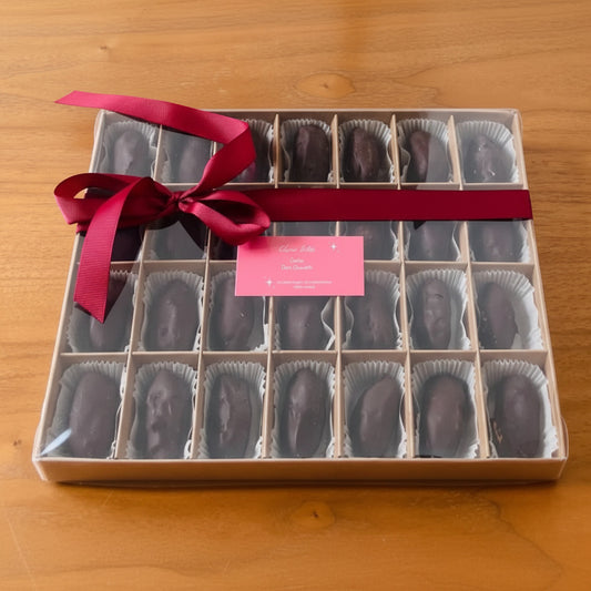 A boxed assortment of chocolates in a clear-lidded tray with 16 compartments, tied with a dark red ribbon and a small pink tag, placed on a wooden surface.
