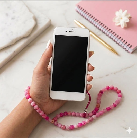 Hand holding a smartphone with a pink beaded strap on a marble surface with stationery items.